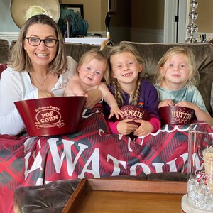 May include: A family of four is gathered on a red and black plaid couch. The mother is holding a baby in her arms. The baby is wearing a white onesie. The two older children are sitting on the couch with their arms around each other. They are all smiling. There are three red wooden bowls with personalized names on them. The bowls say "The Cameron Family's Pop Corn", "Enzie", and "Hutton".