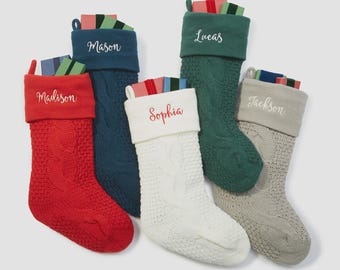 May include: Six cable-knit Christmas stockings in red, navy, green, white, and gray, each with a folded cuff and a name embroidered in a contrasting color: Madison, Mason, Lucas, Sophia, and Jackson. Each stocking is filled with colorful wrapped gifts.