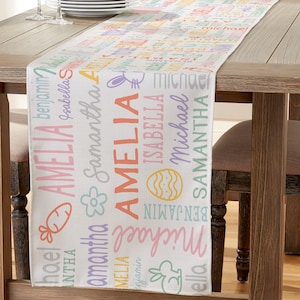 May include: A white table runner with a pastel Easter-themed design. The runner features a repeating pattern of bunnies, eggs, carrots, and flowers. The names Amelia, Samantha, Isabella, Benjamin, and Michael are printed on the runner in a variety of colors.