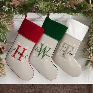 May include: Three personalized Christmas stockings with snowflake designs and the names "Hannah", "William", and "Taylor". The stockings have red, green, and white velvet cuffs.