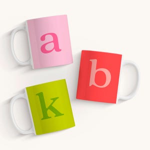 May include: Three ceramic mugs with white handles. The mugs are pink, red, and green. The letter "a" is printed on the pink mug, the letter "b" is printed on the red mug, and the letter "k" is printed on the green mug.