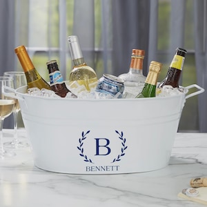 May include: A white metal beverage tub filled with ice and various bottles of wine, beer, and a can. The tub has a navy blue monogram with the letter "B" and the name "BENNETT" below it. The tub has white handles.