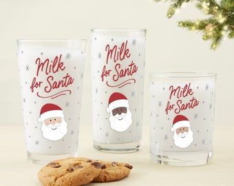 May include: Three glass mugs with a white background and red and black Santa Claus designs. The mugs say "Milk for Santa" in red cursive writing. Two cookies are in the foreground.