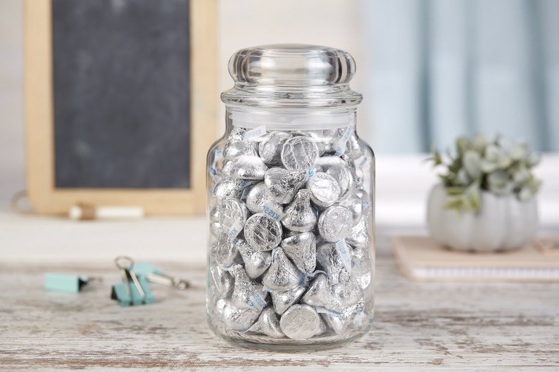 May include: A clear glass jar with a lid filled with silver foil-wrapped chocolate kisses.