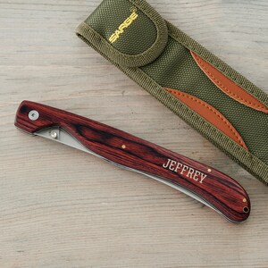 May include: A personalized pocket knife with a wood handle and a stainless steel blade. The knife has a brown leather sheath with a green fabric exterior. The knife is engraved with the name "JEFFREY".