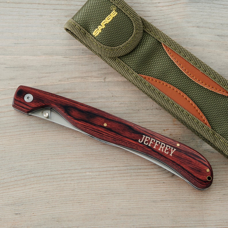 May include: A personalized pocket knife with a wood handle and a stainless steel blade. The knife has a brown leather sheath with a green fabric exterior. The knife is engraved with the name "JEFFREY".