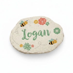 May include: A decorative stone with the name "Logan" in green script. The stone is adorned with colorful flowers and cartoon bees. The design is set against a neutral background, creating a charming garden-themed accent.