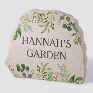 May include: A light-colored, irregular stone plaque featuring the dark brown text "HANNAH'S GARDEN" centered on the front. The stone is decorated with a watercolor-style botanical print of green leaves and small brown berries along the top and bottom edges.