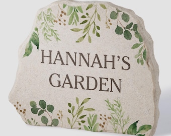 May include: A light-coloured, irregular stone plaque featuring the dark brown text "HANNAH'S GARDEN" centred on the front. The stone is decorated with a watercolour-style botanical print of green leaves and small brown berries along the top and bottom edges.