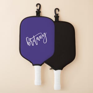 May include: Two purple and black pickleball paddles with white handles. The purple paddle has the name "Brittany" written on it in white cursive.