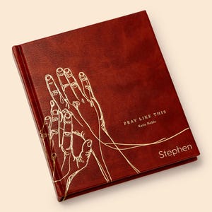 May include: A red leather-bound journal with gold lettering that reads "Pray Like This" by Katie Noble. The journal has a gold illustration of two hands clasped in prayer and the name "Stephen" is printed in gold on the bottom right corner.