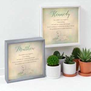 May include: Two framed prints with the names "Matthew" and "Kennedy" in elegant script. Each print features a lamb illustration and a religious verse. The frames are white and gray, with a soft, watercolor-like background.
