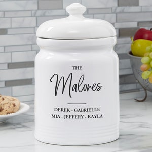 May include: White ceramic cookie jar with black text that reads "The Malores" and "Derek - Gabrielle, Mia - Jeffery - Kayla".