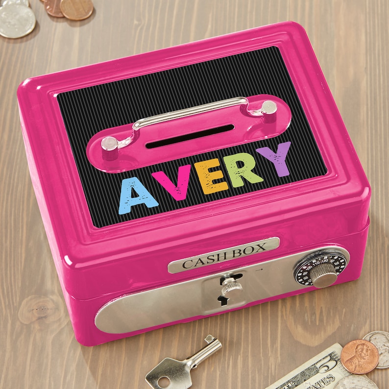 Puede incluir: Una caja registradora rosa brillante con una parte superior negra con el nombre "AVERY" en letras coloridas. La caja tiene un asa plateada, una cerradura de combinaci&oacute;n y las palabras "CASH BOX". Se ve una llave y algunas monedas.
