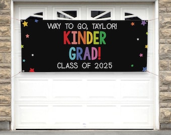 May include: A black banner with the text "WAY TO GO, TAYLOR! KINDER GRAD! CLASS OF 2025" in colorful letters. The banner is decorated with colorful star shapes and is hanging in front of a white garage door.