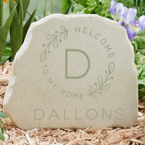 May include: A light beige stone with the words "WELCOME TO MY HOME" and the name "DALLONS" in a soft green. A green "D" is centered within a floral wreath. The stone is set in a bed of wood chips, with greenery and flowers in the background.