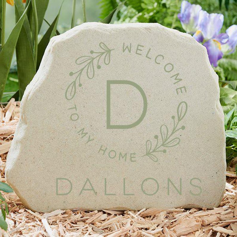 May include: A light beige stone with the words "WELCOME TO MY HOME" and the name "DALLONS" in a soft green. A green "D" is centered within a floral wreath. The stone is set in a bed of wood chips, with greenery and flowers in the background.
