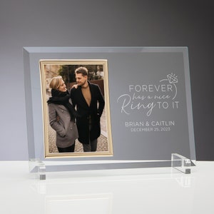 May include: A clear glass photo frame with a gold trim around the photo opening. The frame has a white inscription that reads "FOREVER has a nice Ring TO IT" with a diamond ring symbol. Below the inscription is the text "BRIAN & CAITLIN DECEMBER 25, 2023".