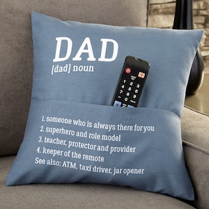 May include: A blue throw pillow with a pocket for a remote control. The pillow has white text that reads "DAD [dad] noun 1. someone who is always there for you 2. superhero and role model 3. teacher, protector and provider 4. keeper of the remote See also: ATM, taxi driver, jar opener"