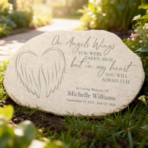 May include: A light-colored stone memorial with engraved text and angel wings. The inscription reads "On Angels Wings...but in my heart...In Loving Memory Of Michelle Williams September 17, 1933 - June 22, 2026."