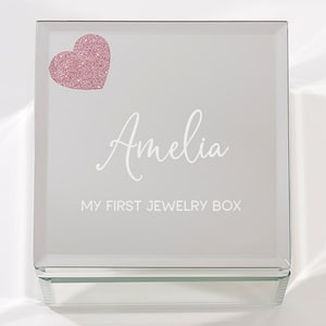 May include: A clear glass jewelry box with a pink glitter heart and the name "Amelia" engraved on the lid. The box also has the text "My First Jewelry Box" engraved on the lid.