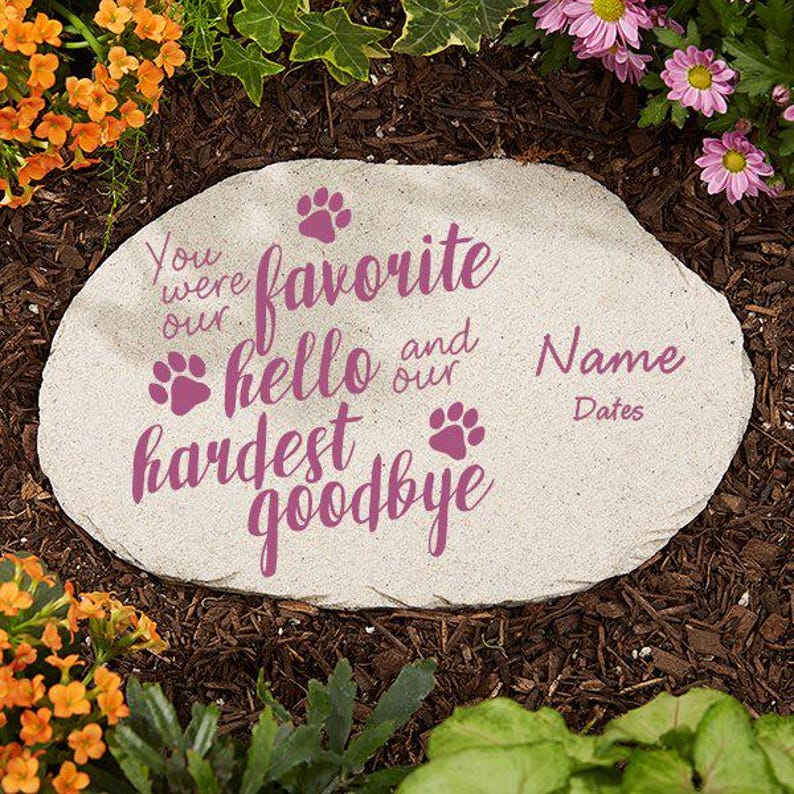 May include: A light-colored stone garden marker with the words "You were our favorite hello and our hardest goodbye" in purple script. Paw prints and space for a name and dates are also included. The stone is surrounded by mulch and flowers.