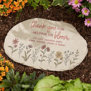 May include: A beige stone garden marker with the words "thank you for helping us bloom" in red script. Below the text are the words "We love Grandma Mary! Emma, Michael, Owen & Sarah". The stone is decorated with floral illustrations.
