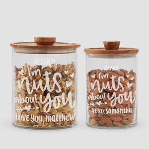 May include: Two clear glass storage jars with wooden lids, filled with nuts. The larger jar features white text reading Jessica, I'm nuts about you, Love you, Matthew. The smaller jar features white text reading Daniel, I'm nuts about you, XOXO, Samantha.