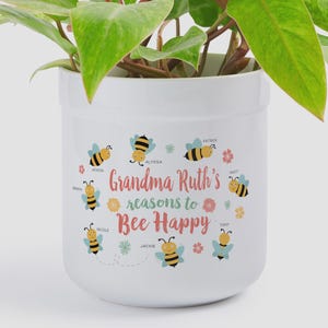 Puede incluir: Una maceta cilíndrica de cerámica blanca con una planta de hojas verdes. La maceta muestra el texto "Grandma Ruth's reasons to Bee Happy" en rosa y verde, rodeado de ilustraciones de seis abejas amarillas y pequeñas flores coloridas.