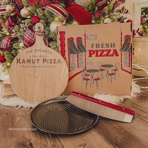 May include: A round wooden pizza serving board engraved with The Rhoades Fresh-Baked Kamut Pizza, a cardboard pizza box with a graphic of tables and chairs, a black circular perforated metal pizza pan, and a red-handled stainless steel pizza cutter.
