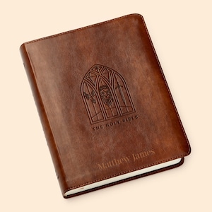 May include: A brown leather-bound Bible with a decorative embossed design of a window with a lion, dove, and sword. The text "The Holy Bible" and "Matthew James" are embossed on the cover.