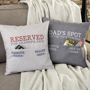 May include: Two gray and white striped throw pillows with pockets. The pillow on the left says "Reserved for Grandpa Jack" and the pillow on the right says "Dad's Spot Don't Get Too Comfortable!" Both pillows have pockets for remotes and snacks.