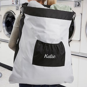 May include: A white and black laundry bag with a black pocket that says "Katie" in white embroidery. The bag is being worn as a backpack.