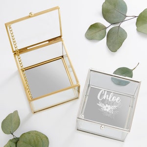 May include: Two glass jewelry boxes with mirrored interiors. One box is gold with a hinged lid and a chain. The other box is silver with a drawer and the name "Chloe" engraved on the front.