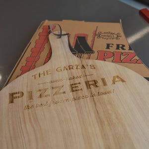 May include: A light-colored wooden pizza peel with a handle and a leather hanging loop. The surface is engraved with the text: THE GARZA'S oven-baked PIZZERIA the best frozen pizza in town! A cardboard pizza box is visible behind the wooden board.