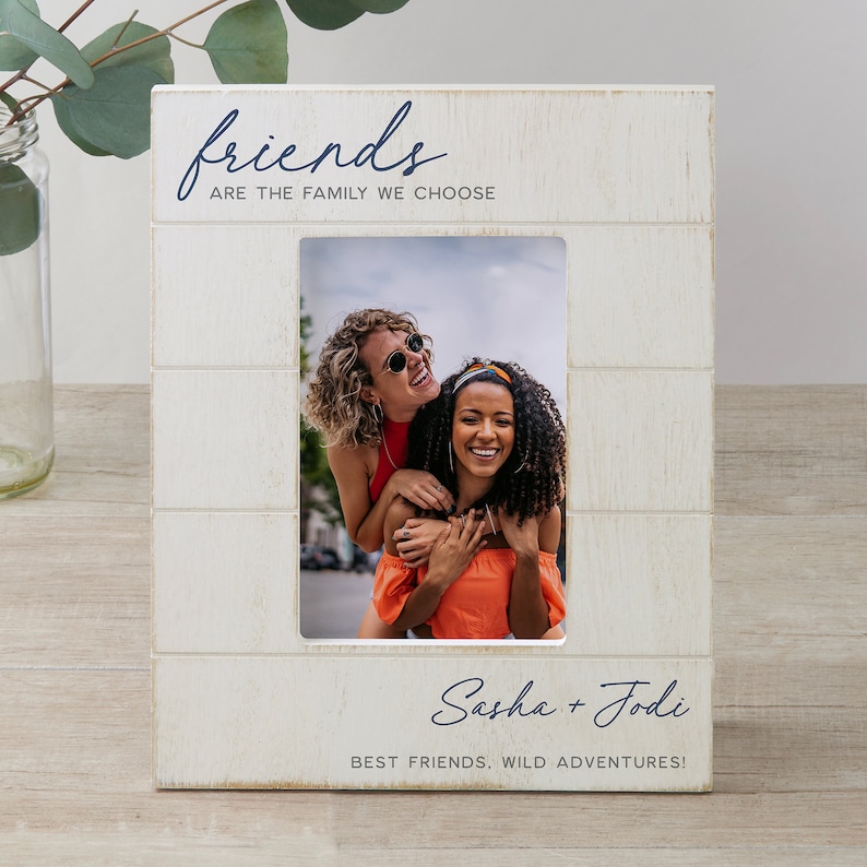 K&ouml;nnte beinhalten: Wei&szlig;er Holzrahmen mit einem wei&szlig;en Passepartout und einem Foto von zwei Freundinnen. Der Rahmen tr&auml;gt den Text "friends are the family we choose" oben und "Sasha + Jodi best friends, wild adventures!" unten.