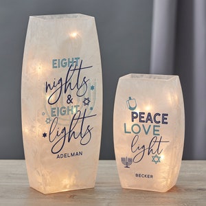 May include: Two white frosted glass vases with LED lights inside. The taller vase has the text "Eight nights & eight lights Adelman" and the shorter vase has the text "Peace Love light Becker".