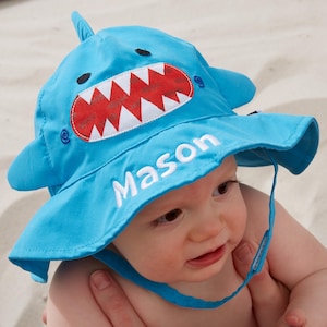 May include: A light blue baby sun hat with a shark design. The hat features a red and white mouth, black eyes, and the name "Mason" embroidered in white. It has a chin strap and a brim for sun protection.