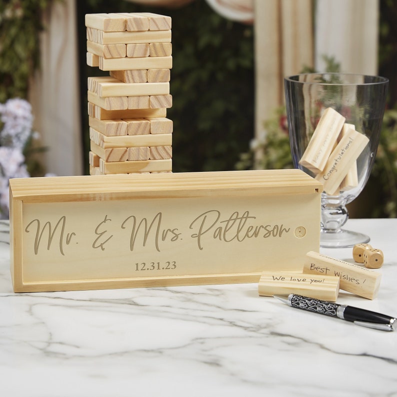 Puede incluir: Un juego de Jenga de madera con una caja personalizada grabada con "Mr. & Mrs. Patterson" y la fecha 12.31.23. Incluye peque&ntilde;os bloques de madera con mensajes como "&iexcl;Felicidades!" y "&iexcl;Mejores deseos!" Tambi&eacute;n hay un bol&iacute;grafo y dados.