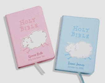 May include: Two Holy Bible books with covers in pastel pink and light blue. Each features a white sheep illustration, a ribbon bookmark, and personalized text. The pink cover reads Gracie Ruth May God Bless You, and the blue reads Issac James October 24, 2026.