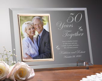 May include: A framed photo with a couple, set against a gray background. The frame is engraved with "50 Years Together" and the names Kathleen and Robert, with the date November 22, 2025. Two roses are in the foreground.