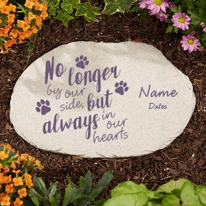 May include: A light-colored stone garden marker with the text "No longer by our side, but always in our hearts" in purple script, along with paw prints. Space is provided for a name and dates. The stone is surrounded by mulch and flowers.