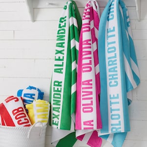 May include: Three beach towels with white stripes and names printed on them. The towels are green, pink, and blue. The names are "Alexander", "Olivia", and "Charlotte".