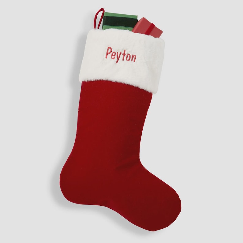 May include: A red Christmas stocking with a white faux fur cuff featuring the embroidered name Peyton in red thread. The stocking has a red hanging loop and contains a small green rectangular gift and a small red rectangular gift inside.