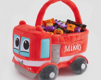 May include: A plush red fire truck basket with a handle, featuring light blue accents, a smiling face, and the name Mikey embroidered in white on the side. The basket is filled with various colorful wrapped candies and a small pumpkin figure.