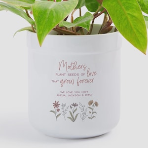 May include: White ceramic plant pot with the text "Mother's love that grow forever". The pot has a floral design and the text "We love you mom! Amelia, Jackson & Emma". The pot contains a green plant.