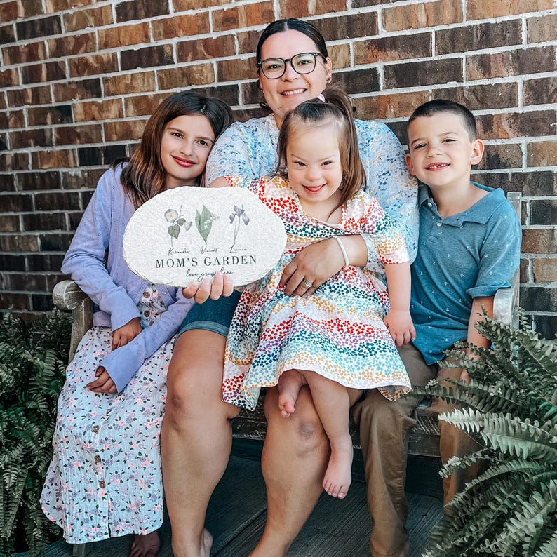 Pu&ograve; includere: Una famiglia di quattro persone &egrave; seduta su una panchina di legno davanti a un muro di mattoni. La madre tiene in mano una pietra decorativa con le parole "MOM'S GARDEN" e illustrazioni floreali. I bambini sorridono e guardano la fotocamera. La pietra &egrave; di forma ovale.