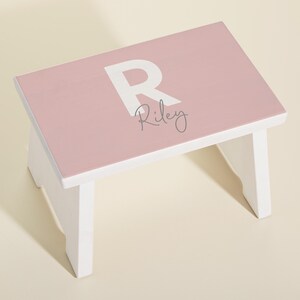 May include: A pink wooden step stool with white legs and a white letter "R" and the name "Riley" painted on the top.