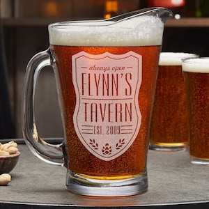 May include: A clear glass beer pitcher with a frosted design. The pitcher is filled with amber beer and has the text "always open - FLYNN'S TAVERN - EST. 2009" etched on the side.