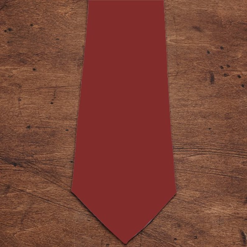 May include: A plain burgundy tie, presented on a wooden surface. The tie is a deep red colour and has a traditional pointed end. The background features a warm-toned wood grain.